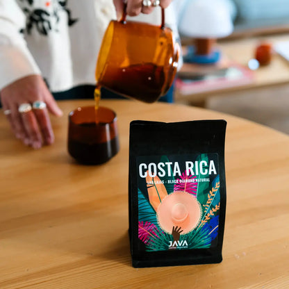 Kawa specialty JAVA Coffee Costa Rica Las Lajas Black Diamond Natural w czarnym opakowaniu na stole, w tle osoba nalewająca kawę z bursztynowego dzbanka do filiżanki