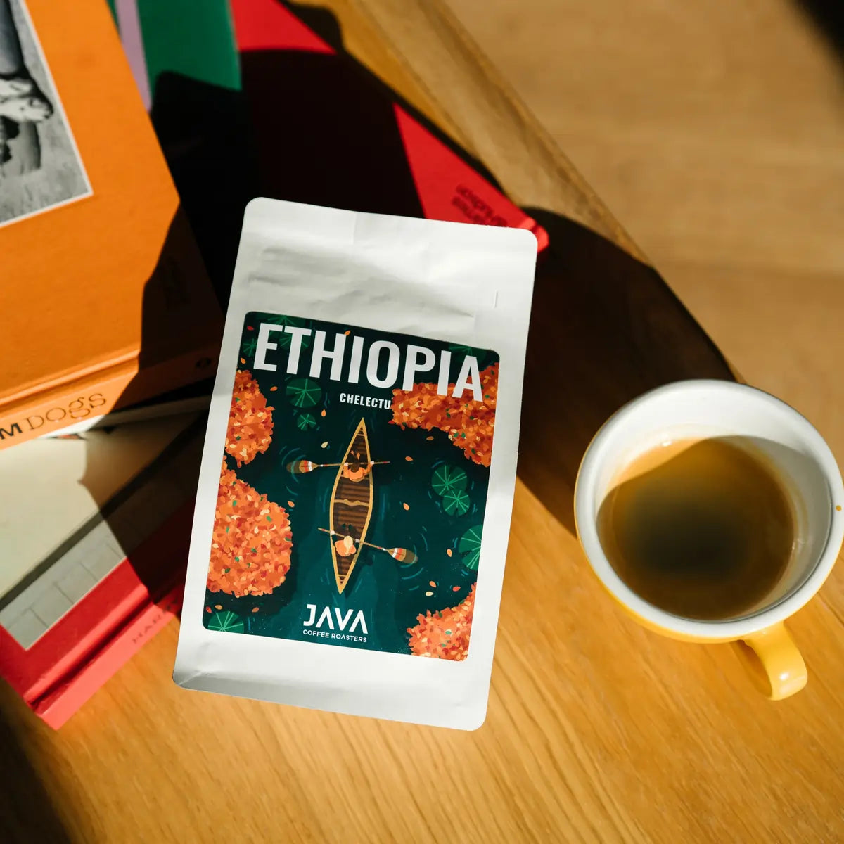 Opakowanie kawy JAVA Coffee Roasters Ethiopia Chelektu leżące na stole obok filiżanki kawy i kolorowych książek w promieniach słońca.