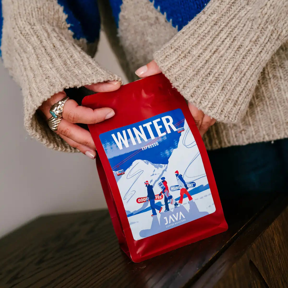 Osoba trzyma czerwone opakowanie kawy JAVA Coffee Winter Espresso, ustawiając je na stole w domowym wnętrzu