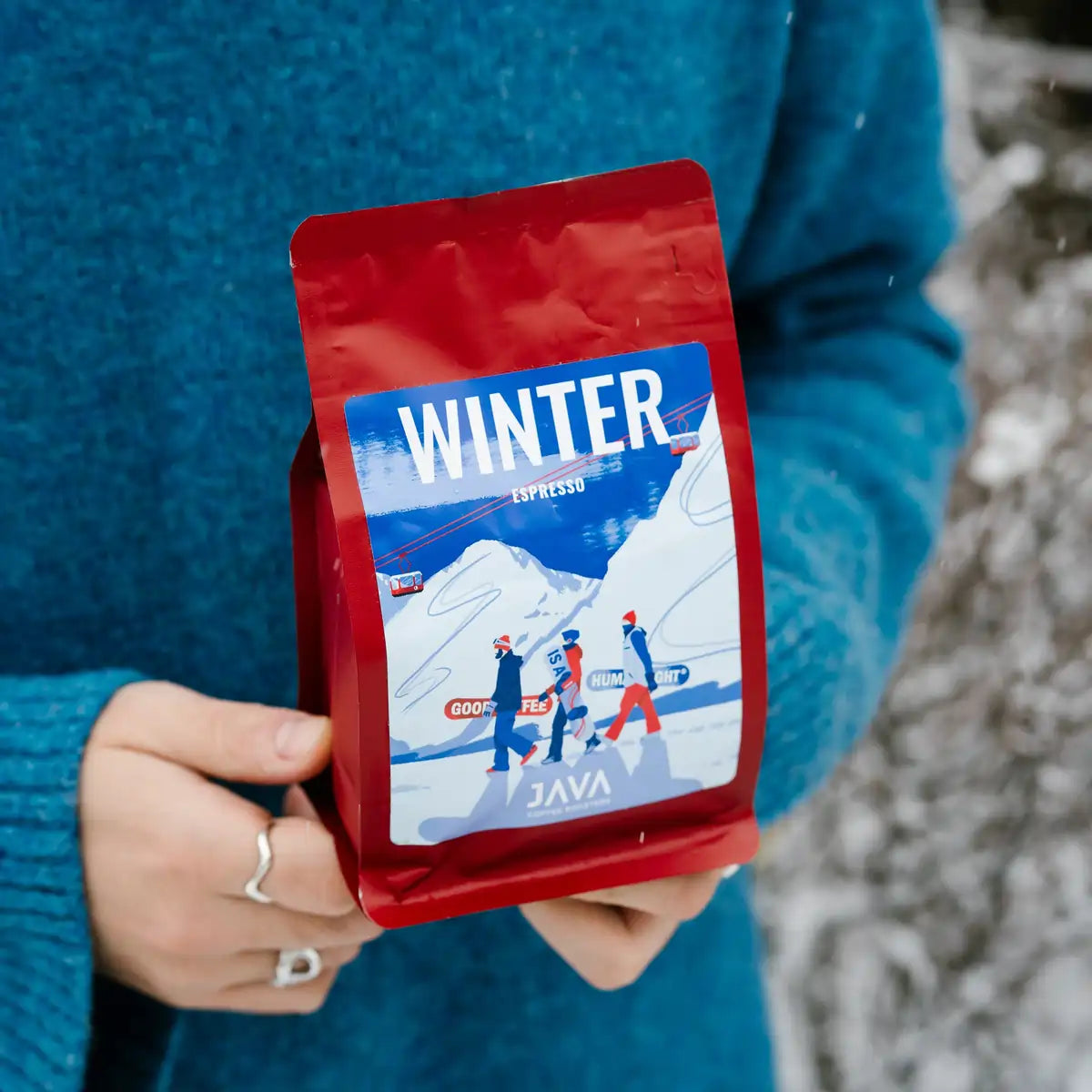Osoba trzyma w dłoniach czerwone opakowanie kawy JAVA Coffee Winter Espresso w zimowym otoczeniu.
