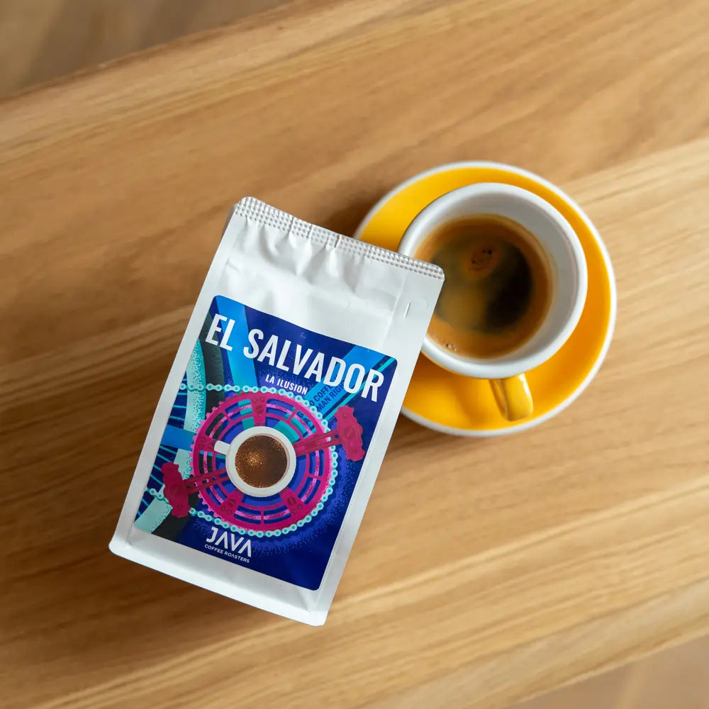 Filiżanka espresso w żółtym spodku obok paczki kawy ziarnistej El Salvador La Ilusion od Java Coffee Roasters, leżącej na drewnianym stoliku.