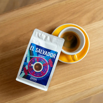 Filiżanka espresso w żółtym spodku obok paczki kawy ziarnistej El Salvador La Ilusion od Java Coffee Roasters, leżącej na drewnianym stoliku.