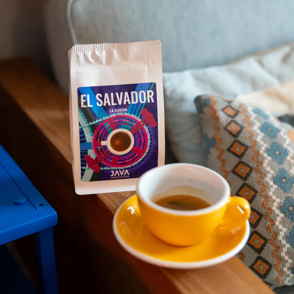Filiżanka espresso w żółtym spodku obok paczki kawy ziarnistej El Salvador La Ilusion od Java Coffee Roasters, leżącej na drewnianym stoliku obok książki i poduszki z etnicznym wzorem.