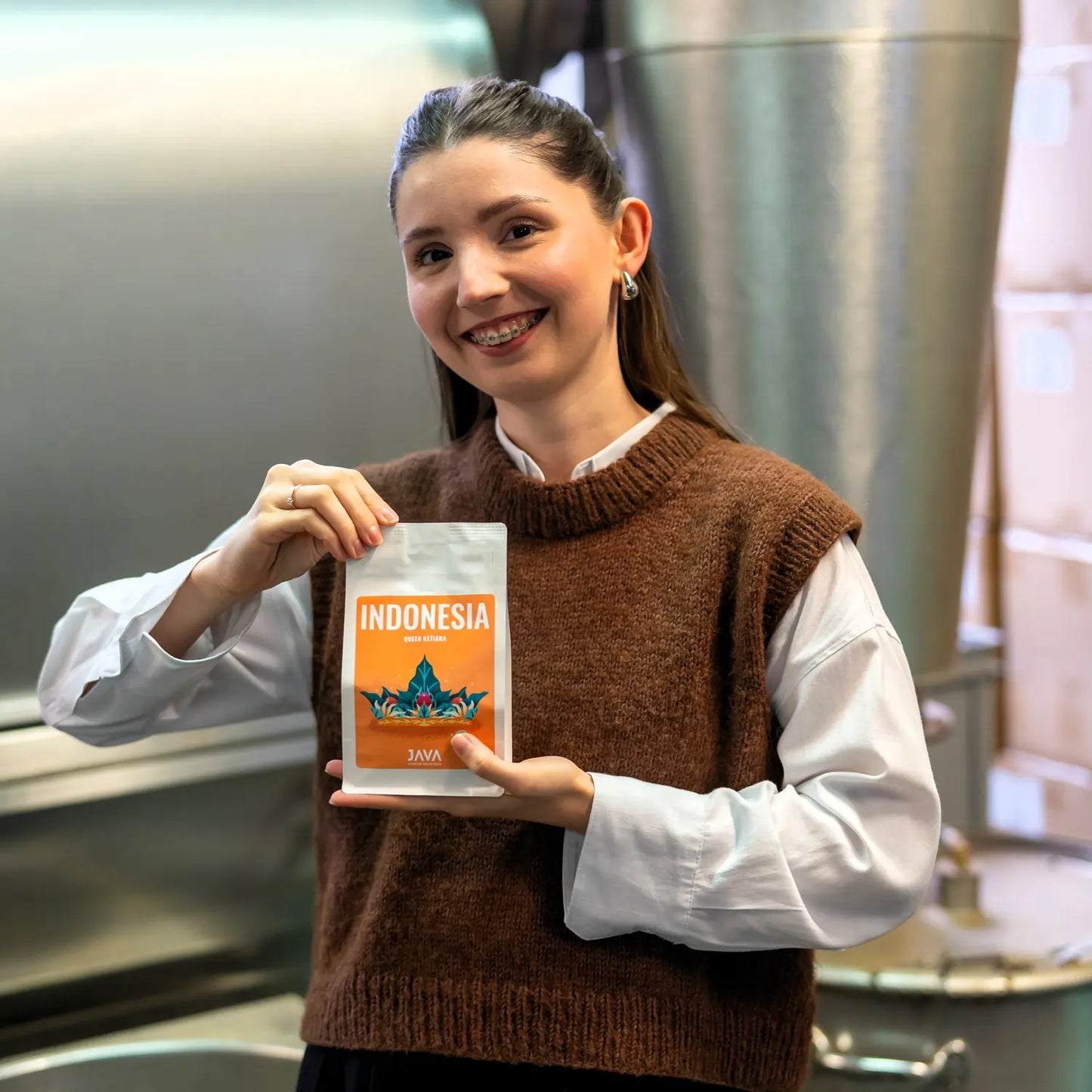 Pracowniczka palarni JAVA Coffee Roasters trzymająca opakowanie kawy Indonesia Queen Ketiara na tle przemysłowych zbiorników w palarni.