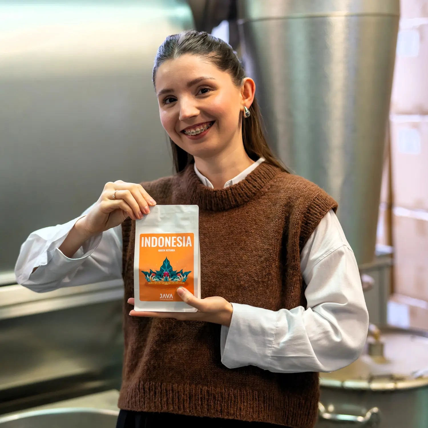 Pracowniczka palarni JAVA Coffee Roasters trzymająca opakowanie kawy Indonesia Queen Ketiara na tle przemysłowych zbiorników w palarni.