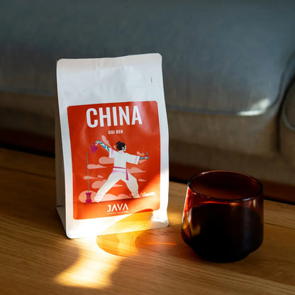 Opakowanie kawy JAVA Coffee Roasters China Gui Ben stojące na drewnianym stole w promieniach słońca obok filiżanki z czarną kawą.