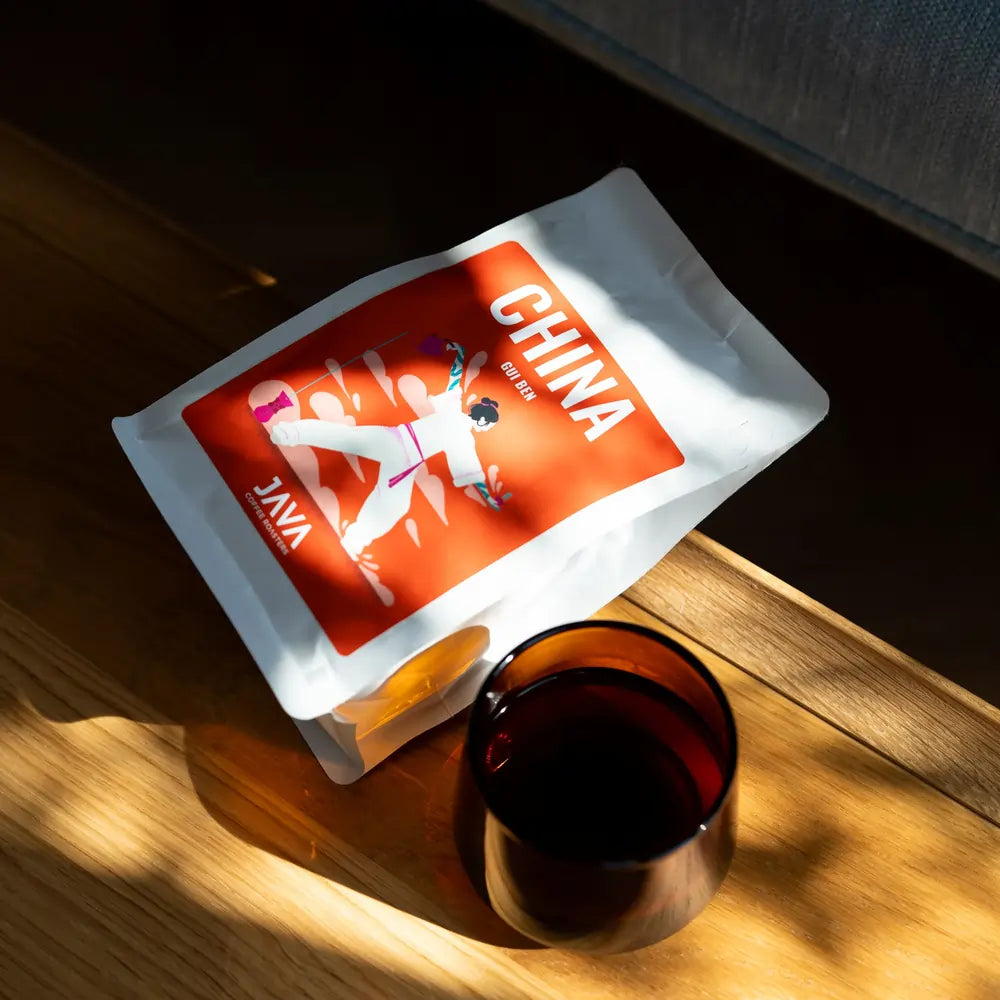 Opakowanie kawy JAVA Coffee Roasters China Gui Ben na drewnianym stole w promieniach słońca obok szklanki czarnej kawy.