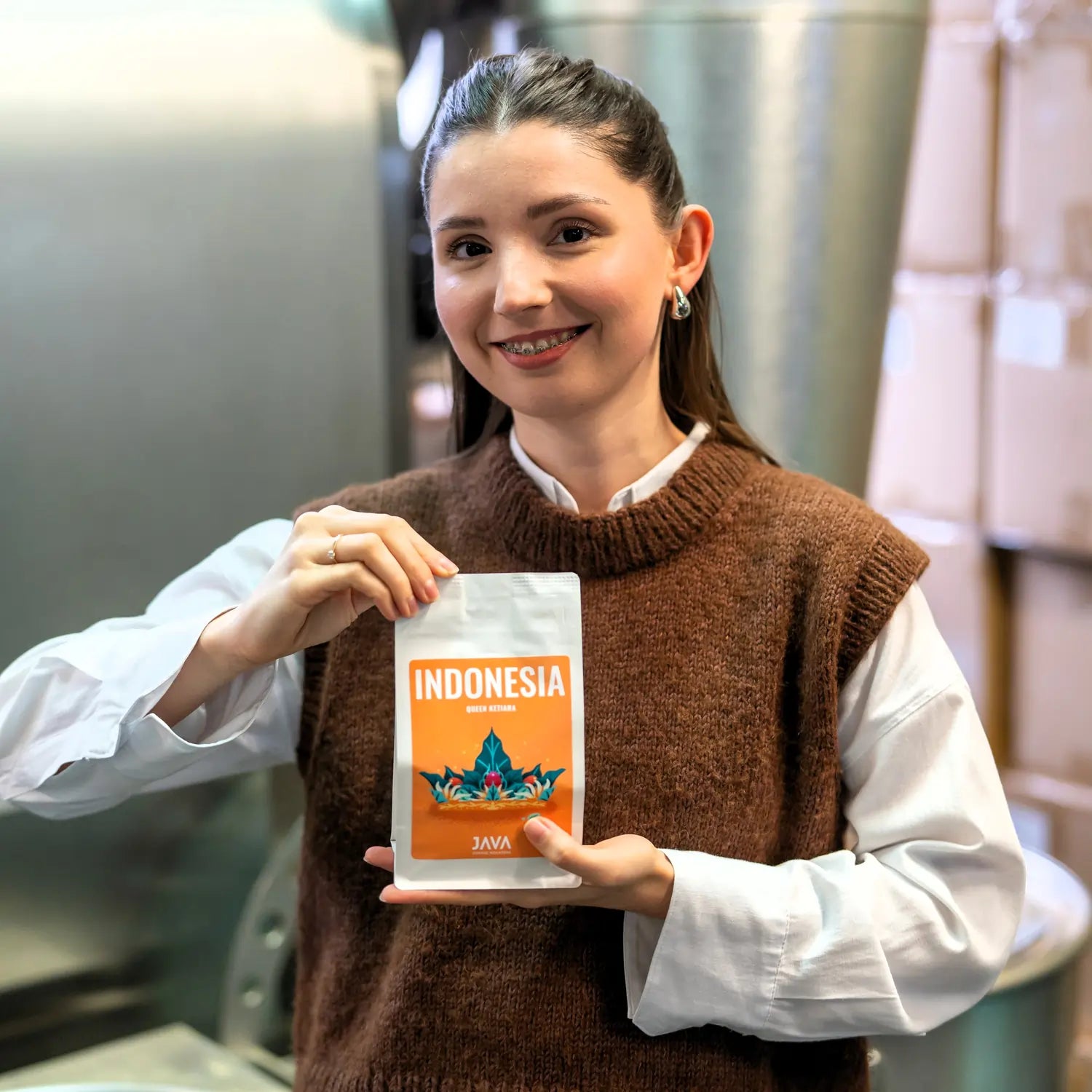 Baristka JAVA Coffee Roasters prezentująca kawę specialty Indonesia Queen Ketiara na tle palarni i kartonów z kawą.