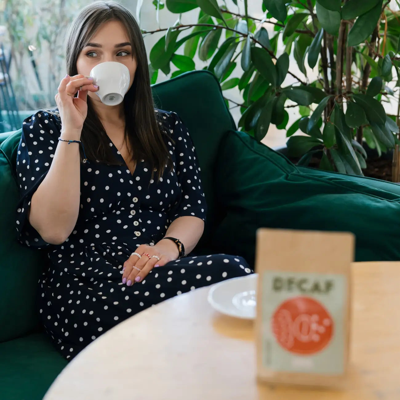 Kobieta w sukience w groszki pije kawę bezkofeinową JAVA Decaf, siedząc na zielonej sofie; w tle widoczna paczka kawy na stole