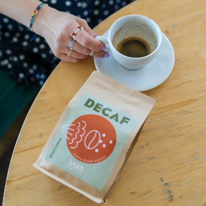  Zbliżenie na filiżankę espresso i paczkę bezkofeinowej kawy specialty JAVA Decaf Kenya Kijani Kiboko na drewnianym stole – ręka z kolorowymi pierścionkami trzyma filiżankę