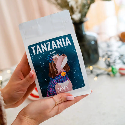 Zbliżenie na opakowanie kawy JAVA Coffee Tanzania Itumpi trzymane w dłoniach w świątecznym, przytulnym wnętrzu.