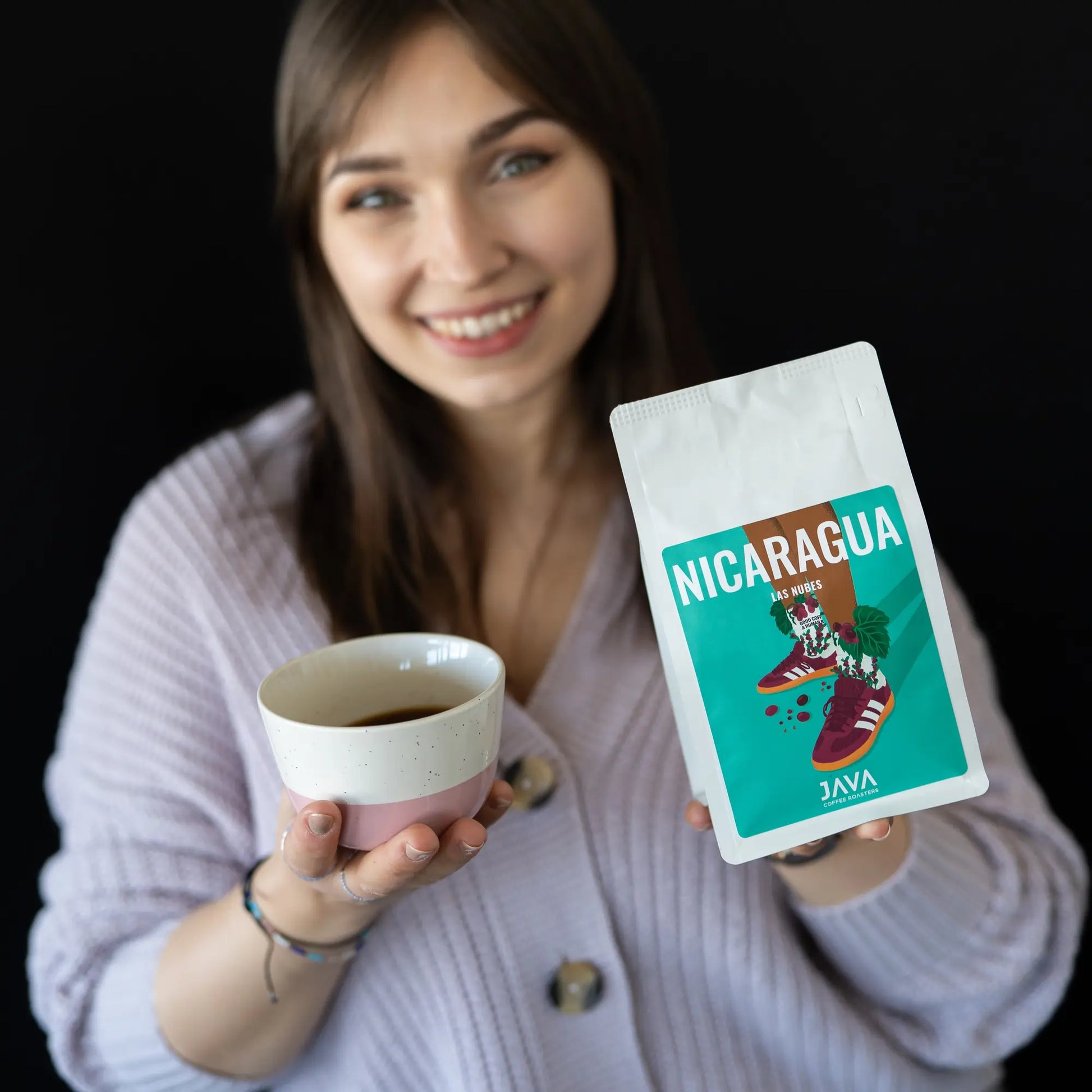 Kobieta w jasnym swetrze uśmiecha się i trzyma w jednej ręce filiżankę z czarną kawą, a w drugiej opakowanie kawy Nicaragua Las Nubes od JAVA Coffee Roasters. Na opakowaniu widnieje ilustracja przedstawiająca buty ozdobione liśćmi i owocami kawowca na czarnym tle.
