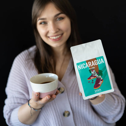 Kobieta w jasnym swetrze uśmiecha się i trzyma w jednej ręce filiżankę z czarną kawą, a w drugiej opakowanie kawy Nicaragua Las Nubes od JAVA Coffee Roasters. Na opakowaniu widnieje ilustracja przedstawiająca buty ozdobione liśćmi i owocami kawowca na czarnym tle.