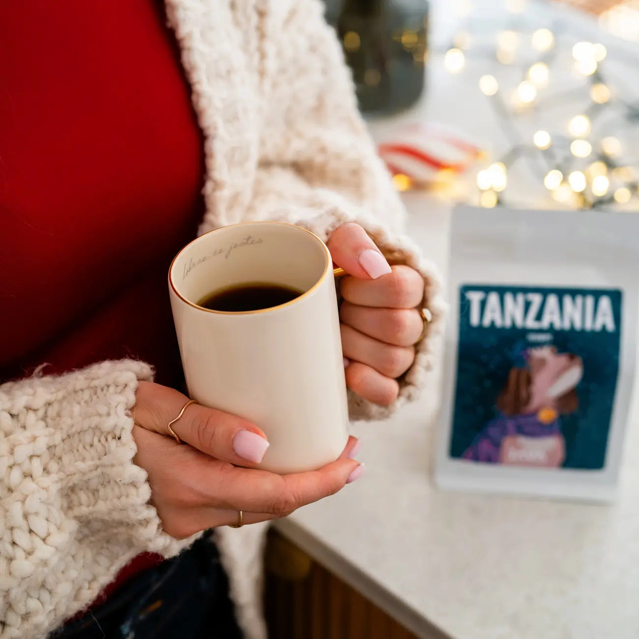 Kobieta trzyma kubek z kawą, a w tle stoi opakowanie JAVA Coffee Tanzania Itumpi w świątecznym wnętrzu.
