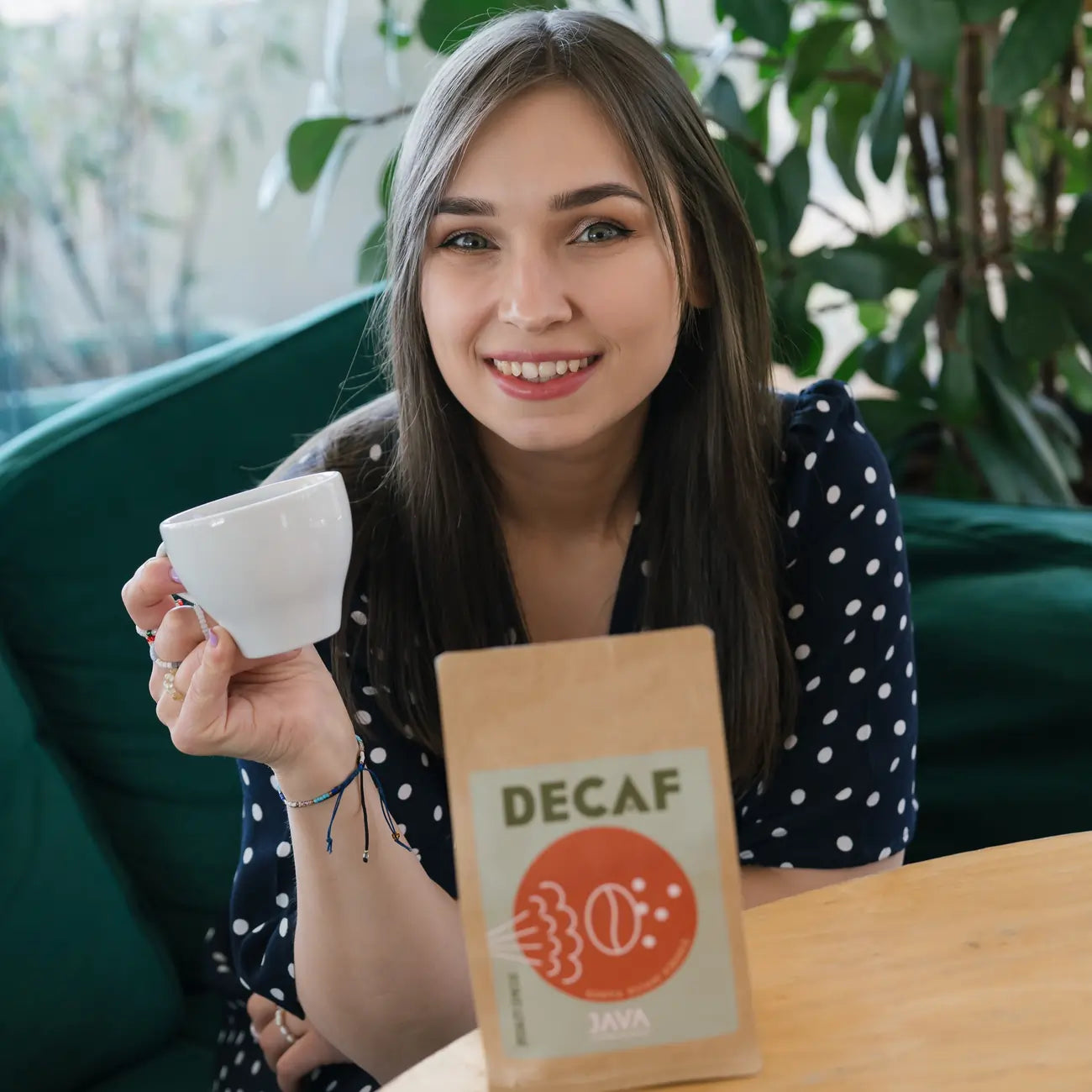 Kobieta z kubkiem kawy uśmiecha się do kamery; na pierwszym planie widoczna paczka kawy bezkofeinowej JAVA Decaf na stole