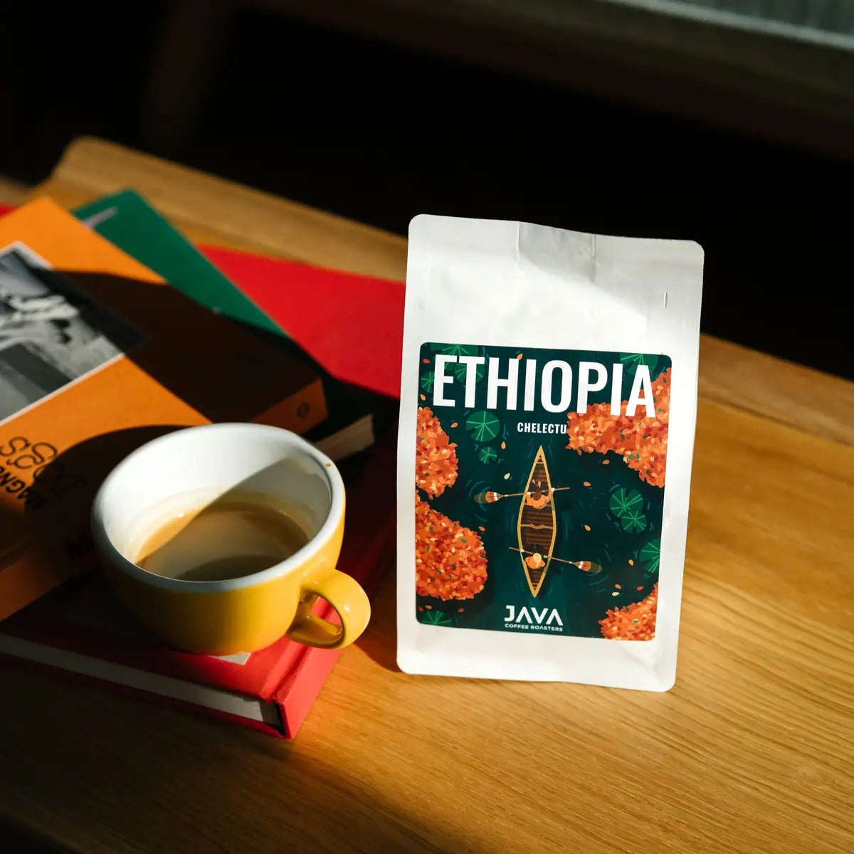  Paczka kawy JAVA Coffee Roasters Ethiopia Chelektu stojąca na drewnianym stole obok filiżanki espresso i książek w ciepłym świetle dziennym.