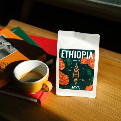  Paczka kawy JAVA Coffee Roasters Ethiopia Chelektu stojąca na drewnianym stole obok filiżanki espresso i książek w ciepłym świetle dziennym.