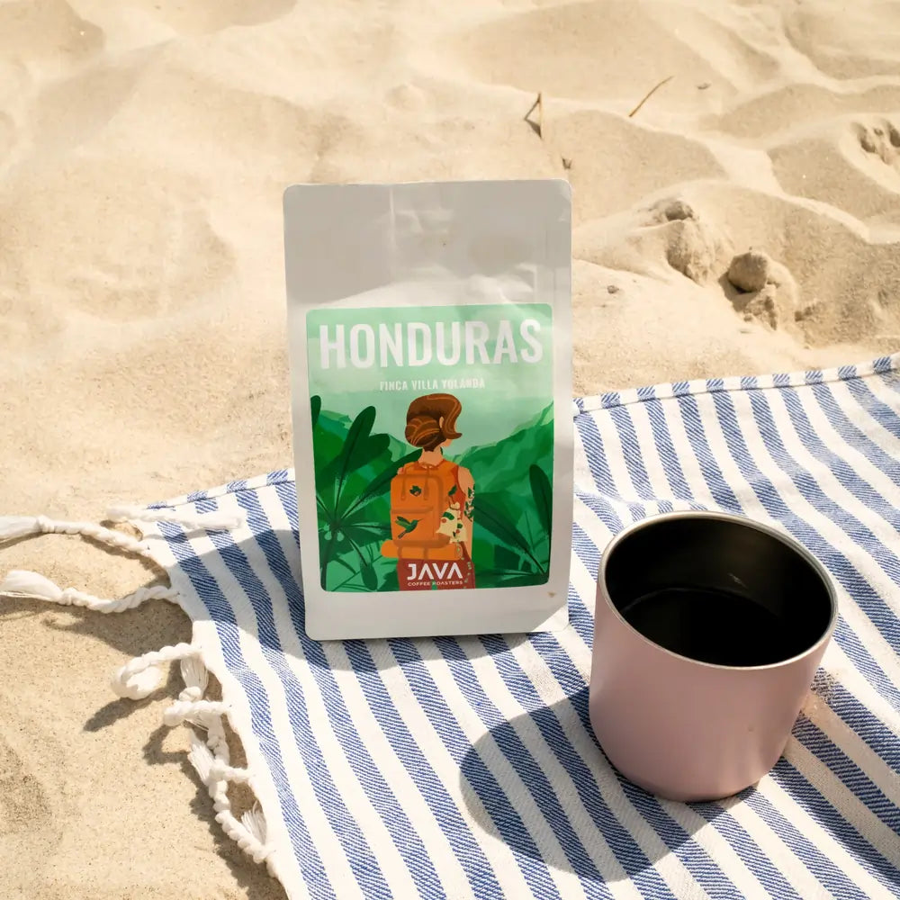 Kawa ziarnista Honduras Finca Villa Yolanda 250 g JAVA Coffee Roasters na plaży – opakowanie na ręczniku w paski obok metalowego kubka, sceneria lifestyle