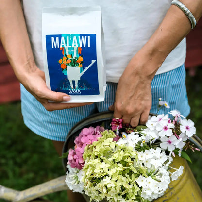 Opakowanie kawy Malawi Ngapani od JAVA Coffee Roasters trzymane w dłoni obok żółtej konewki z kolorowymi kwiatami – oferta kawy specialty dla biur i gastronomii.