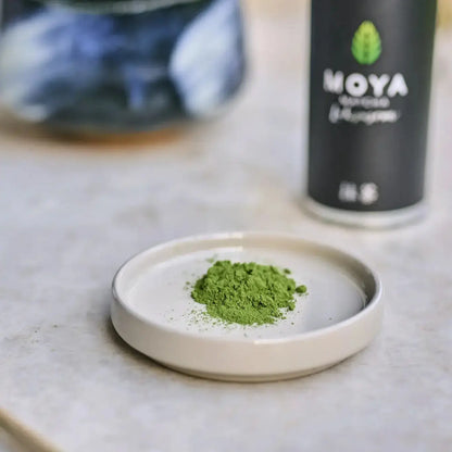 Zbliżenie na sproszkowaną herbatę Moya Matcha Luksusowa na ceramicznym spodku – intensywnie zielony proszek w tle z opakowaniem herbaty.