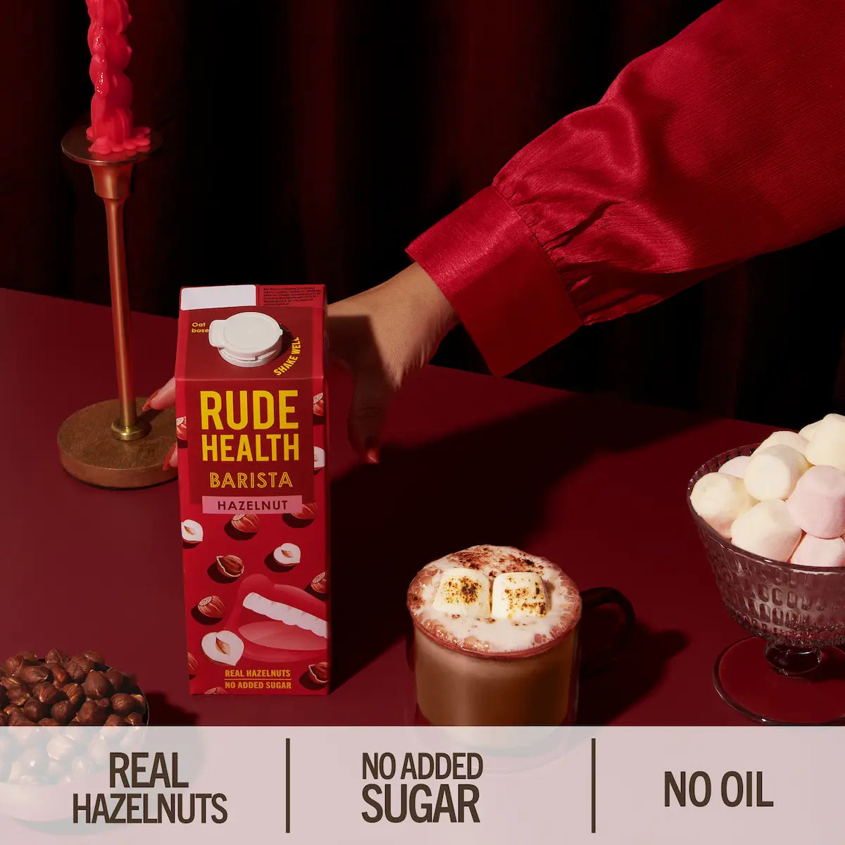 Napój roślinny Rude Health Barista Hazelnut 1L na bordowym tle, obok filiżanki gorącej czekolady z piankami i miseczki orzechów laskowych, podkreślający naturalny skład – bez cukru i oleju.