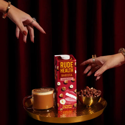 Napój roślinny Rude Health Barista Hazelnut 1L na złotej tacy obok filiżanki kawy i miseczki z orzechami laskowymi, na tle bordowych zasłon, w eleganckiej stylizacji.
