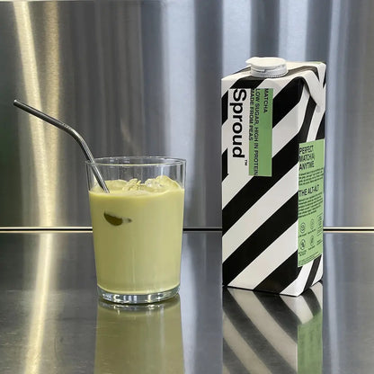 Szklanka z napojem roślinnym Sproud Matcha na lodzie obok kartonu napoju na stalowym blacie