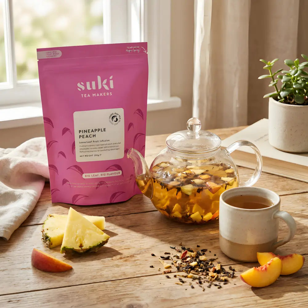 Herbata owocowa SUKI Tea Pineapple Peach – opakowanie, dzbanek z naparem i filiżanka herbaty na drewnianym stole